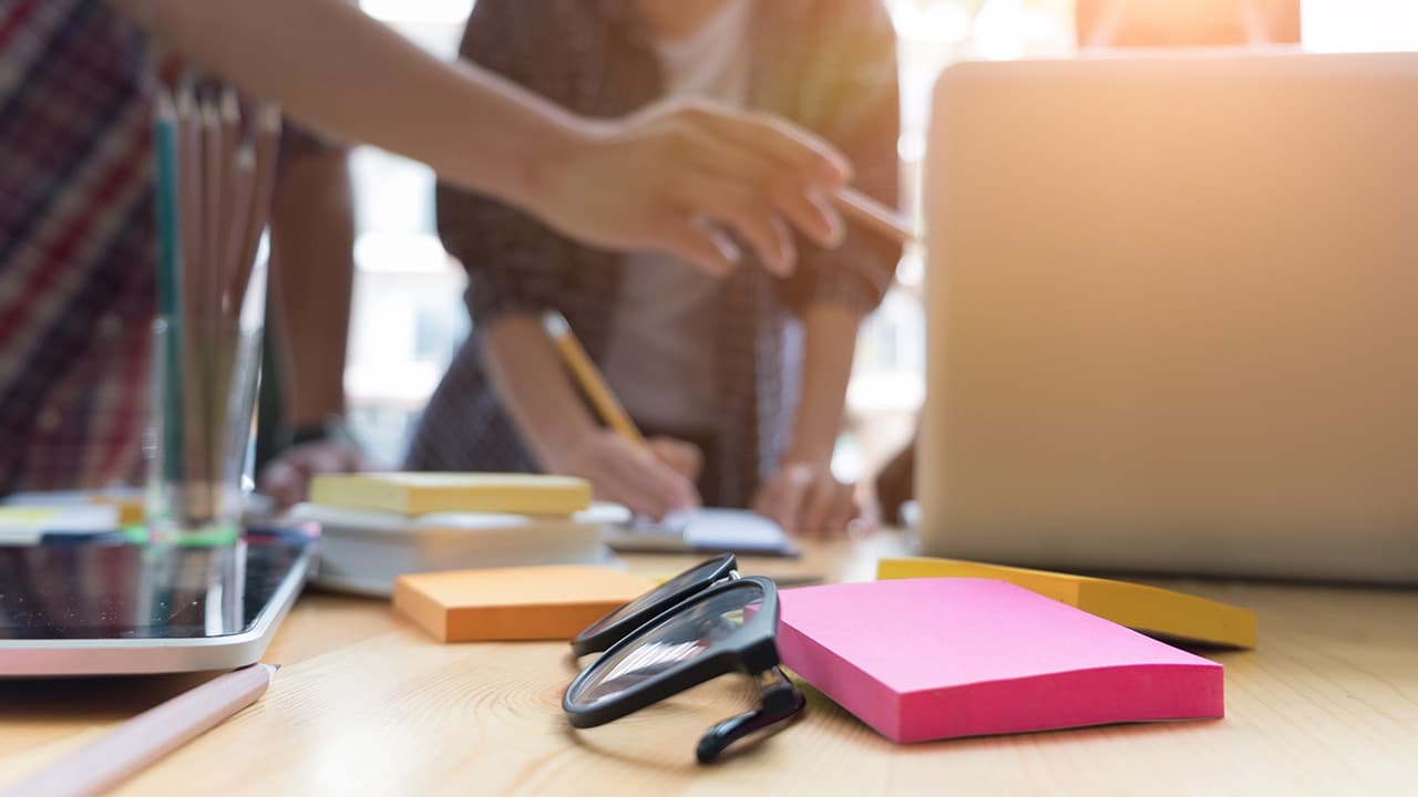 Íróasztal fókuszban lévő szemüveggel és színes post-it jegyzetekkel; a háttérben elmosódva diákok dolgoznak közösen.