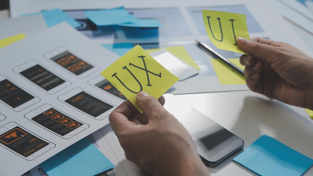 Weboldal tervezési folyamat, ahol a szakember sárga UX és UI feliratú post-it jegyzeteket tart a kezében egy mobilalkalmazás vázlata felett.