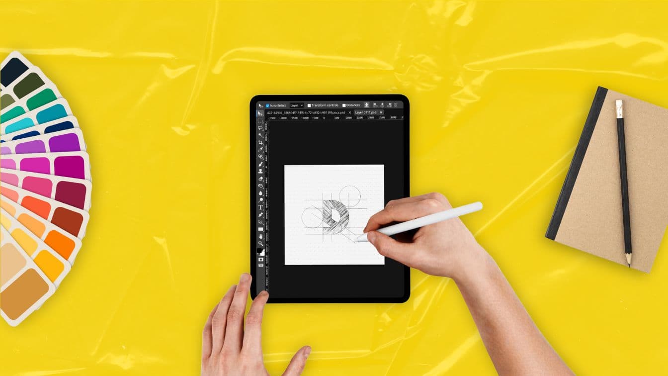 Felülnézetből egy kéz logót tervez tableten digitális tollal, mellette színes színminta és egy füzet sárga háttéren.