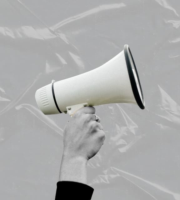 A hand holding up a white megaphone against a textured, crinkled grey plastic background.