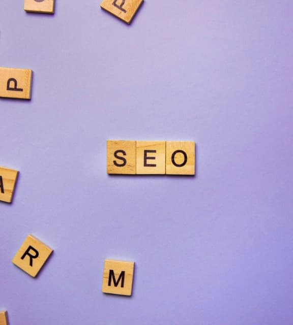 Wooden letter tiles spelling "SEO" in the center of a light purple background with other scattered letters.